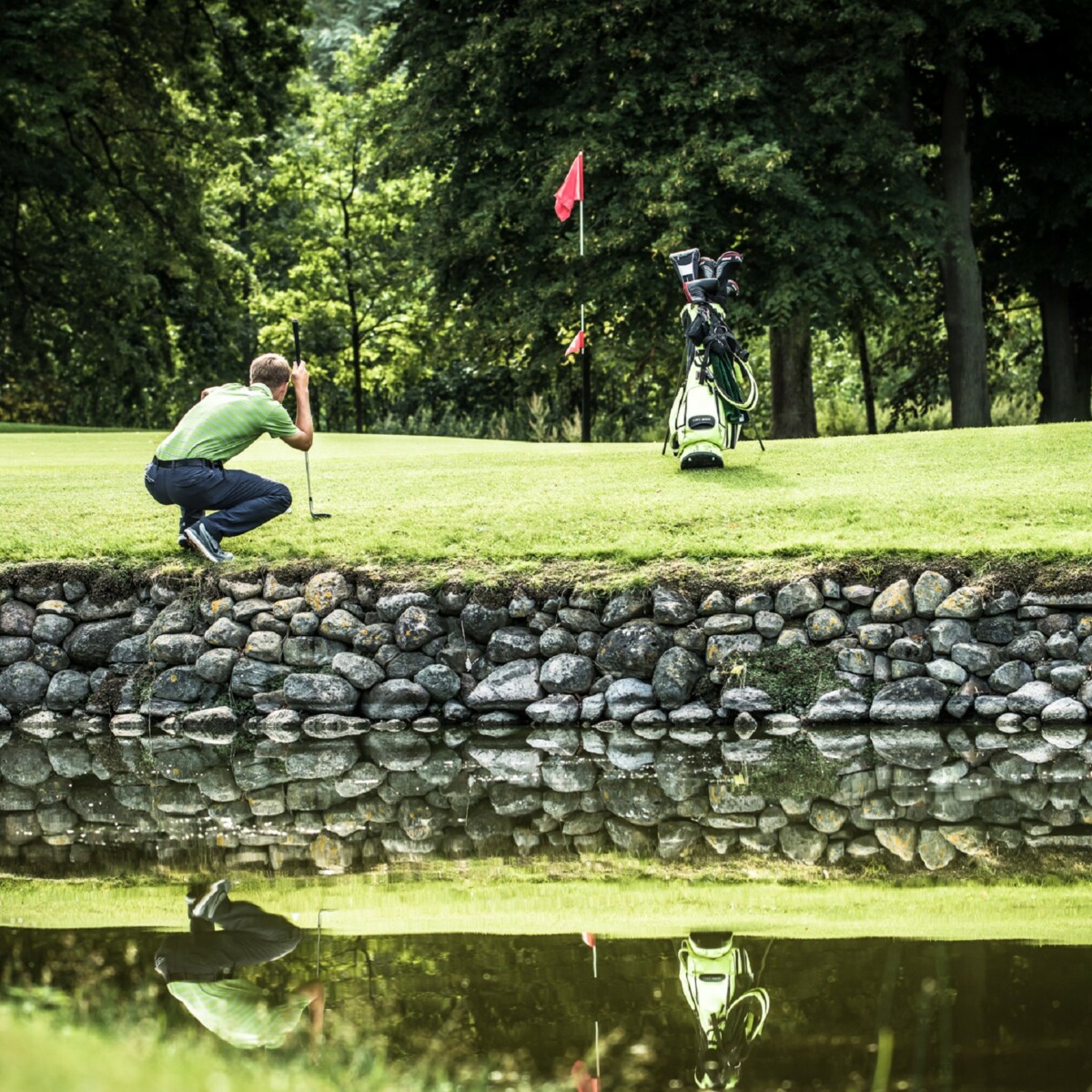 Ein Mann kniet auf einem Golfplatz am Rand des Ufers vor einem Teich.