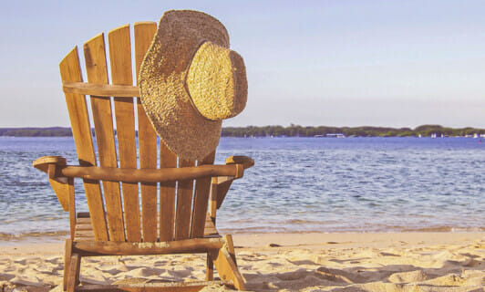 Ein Strandstuhl aus Holz am See Strand, auf dem ein Strohhut hängt.