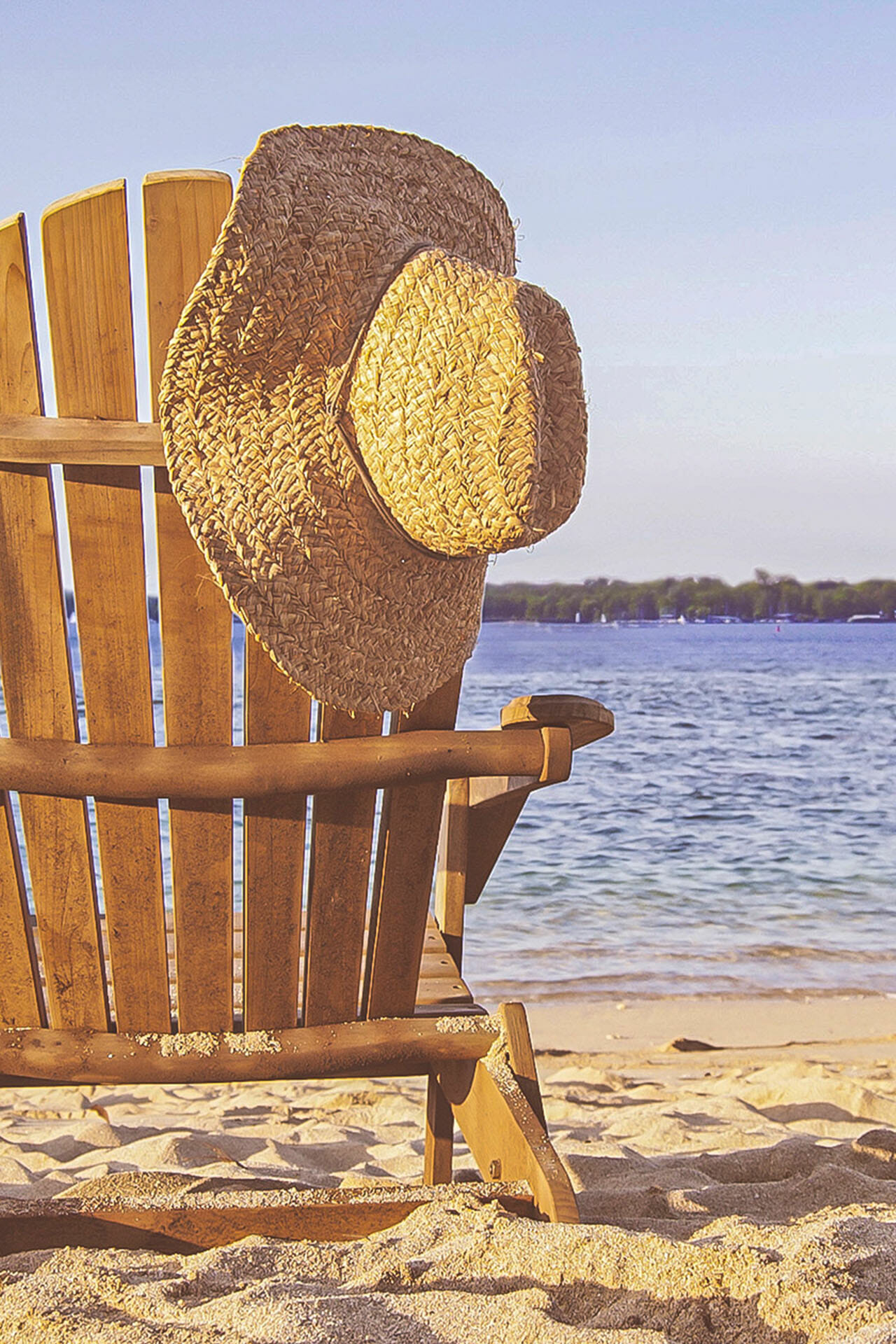 Ein Strandstuhl aus Holz am See Strand, auf dem ein Strohhut hängt.