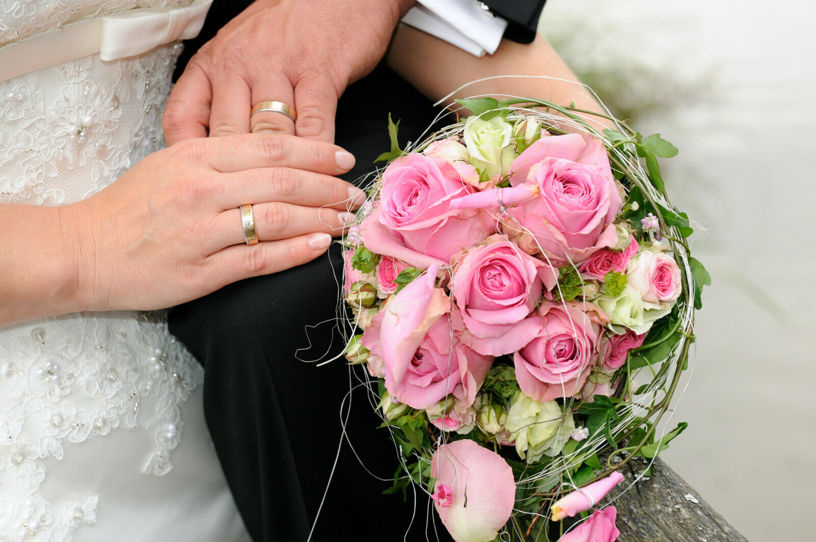 Hände mit Eheringen und ein rosa Blumenstrauß.